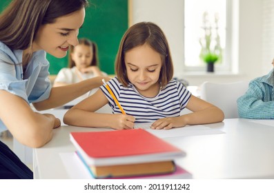 Happy Teacher Helping Her Students In Elementary School. Close Up Portrait Of A Little Schoolgirl And A Female Teacher Who Helps To Complete The Task Correctly. Concept Of Education And Teaching.