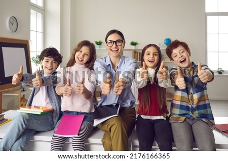 Happy teacher with pupils group giving thumbs up. Toothy smiling tutor with elementary or secondary school student looking at camera portrait. Schoolroom interior. Offline education acknowledgment