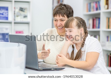Happy teacher and smiling girl with down syndrome use a laptop at library. Education for disabled children concept