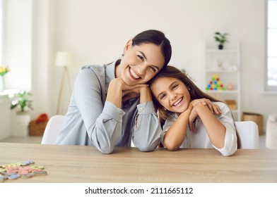 Happy Teacher And Student Girl Sitting At Working Desk In Classroom. Cheerful Young Mother And Child Sitting At Table And Smiling. Little Kid And Her Private Language Tutor Looking At Camera Together