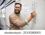 Happy teacher teaching how to do the column additions at elementary school. Smiling man explaining additions in column in primary class while writing on whiteboard. Math’s teacher explaining sums.