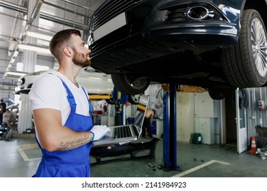 Happy technician mechanic man 20s wear blue overalls t-shirt use hold laptop pc computer stand near car lift check technical condition work in vehicle repair shop workshop indoors Tattoo translate fun