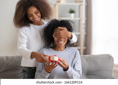 Happy teen african american girl closing mum eyes make surprise to excited black mommy giving birthday gift box congratulating mixed race mom with mothers day holding present sitting on sofa at home