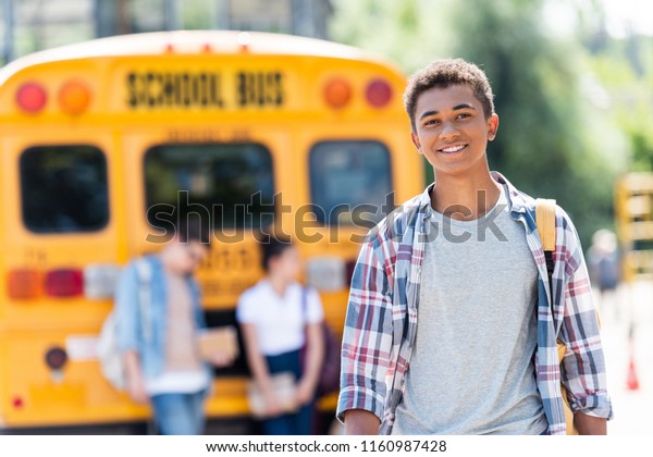 happy teen african american schoolboy looking\
at camera in front of school\
bus