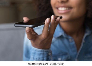 Happy teen African girl giving voice commands to virtual assistant on smartphone, speaking for recording audio message, holding, using mobile phone, smiling. Hand with cellphone close up
