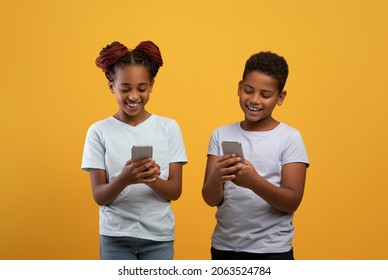 Happy teen black brother and sister playing games, using social media or checking new mobile applications on smartphone, having conversation, yellow studio background. Gadget addiction among kids