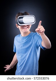 Happy Teen Boy Wearing Virtual Reality Goggles Playing Video Games, On Black Background. Cheerful Smiling Looking In VR Glasses And Gesturing With His Hand. Child Experiencing 3D Gadget Technology.