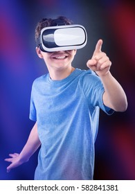 Happy Teen Boy Wearing Virtual Reality Goggles Playing Video Games, On Colorful Bokeh Background. Cheerful Smiling Looking In VR Glasses And Gesturing With His Hand. Child Experiencing 3D Gadget