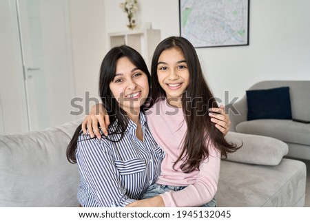 Happy teen child daughter hugging young indian mother at home, portrait. Smiling ethnic family mom with teenage kid girl embracing, bonding together looking at camera sitting on couch.