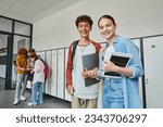 happy teen classmates holding devices and looking at camera in school hallway, adolescent students