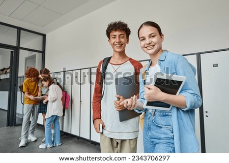 happy teen classmates holding devices and looking at camera in school hallway, adolescent students