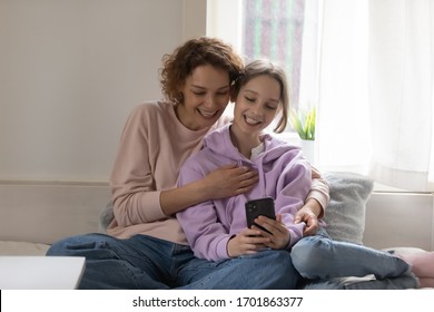 Happy Teen Daughter And Young Mum Using Smartphone At Home Taking Selfie, Watching Funny Social Media Video Bonding At Home. Smiling Parent Mother Hugs Teenager Child Having Fun Playing Mobile Game.