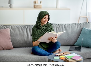 Happy Teen Girl In Hijab Sitting On Sofa With Textbook, Reading, Studying Online From Home, Preparing For Exam. Smart Eastern Adolescent In Headscarf Learning Remotely During Coronavirus Lockdown