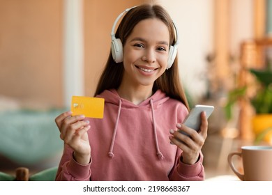 Happy Teen Girl Holding Credit Card And Paying With Smartphone, Listening Music In Wireless Headphones, Sitting At Home. Shopaholic, Online Shopping In Store, App For Buying Purchases