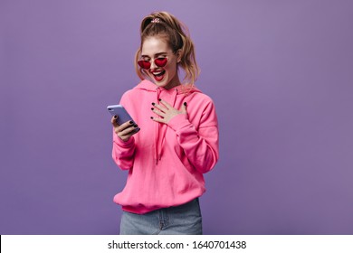 Happy teen girl in hoodie reading message and smiling on purple background. Portrait of pretty young woman in denim skirt and red sunglasses holding cell phone