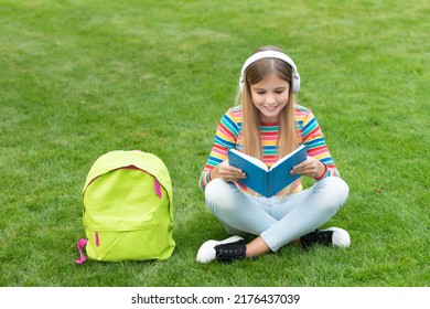 Happy Teen Girl Reading And Listening To Audiobook In Headphones Sitting On Grass Outdoors, School