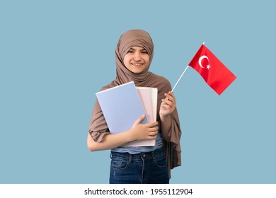Happy Teen Girl In Traditional Muslim Hijab Holding Turkish Flag And Textbooks On Blue Studio Background. Lovely Teenager Lady In Headscarf Posing And Smiling. Education For Arab Females Concept