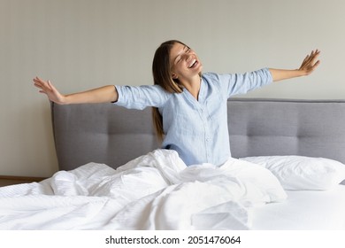 Happy teen millennial girl feeling joy, full of energy after sleep enough, awaking. Cheerful young woman sitting in bed on comfortable mattress, stretching hands, body, smiling with closed eyes - Shutterstock ID 2051476064
