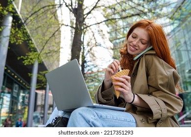 Happy Teen Redhead Hipster Girl Student Using Laptop, Taking On The Phone While Eating Ice Cream In City, Online Learning Outdoors, Sitting On Urban Street Spending Time Outside.