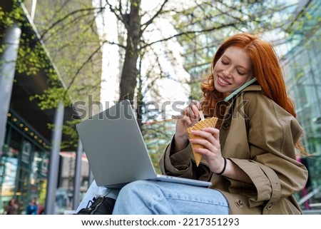 Happy teen redhead hipster girl student using laptop, taking on the phone while eating ice cream in city, online learning outdoors, sitting on urban street spending time outside.