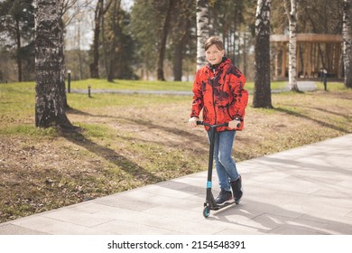 Happy Teen Riding Scooter In Park. Ecological Urban Transport. Caucasian Teenager Boy In Red Jacket Having Fun Driving Scooter Through Urban City Street On Spring Day. Child Walking Outdoors.