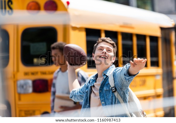 happy\
teen schoolboy throwing american football ball in front of school\
bus with blurred classmate walking on\
background