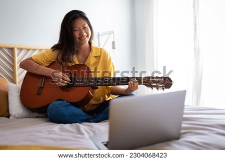 Happy teenage asian girl learning to play guitar with online teacher using laptop in bedroom sitting on bed. Copy space.
