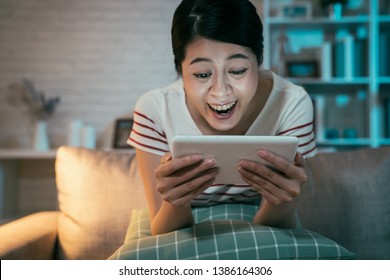 Happy Teenage Asian Girl Sitting On Couch At Home Enjoy Funny Movie On Tablet In Dark Living Room. Young Woman Laughing On Comedy Talk Show On Internet On Digital Pad. Binge Watching Lifestyle Night