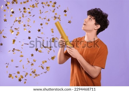 Happy teenage boy blowing up party popper on violet background