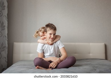 Happy Teenage Boy Sitting Home On Bed, Little Girl, Brother And Sister Hugging Him From Behind. Concept Happy Family, Childhood