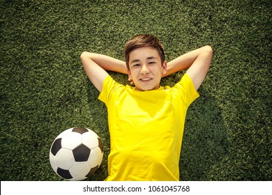 Happy Teenage Boy With A Soccer Ball Lying On The Green Lawn. Shot From Top.