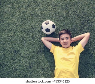 Happy Teenage Boy With A Soccer Ball Lying On The Green Lawn. Shot From Top.