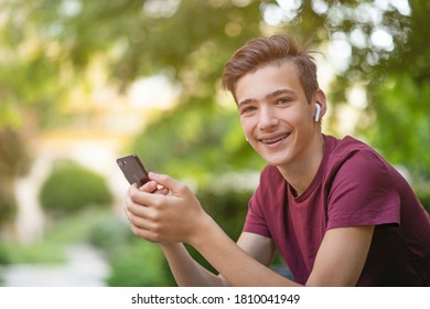 Happy Teenage Boy Is Using Mobile Phone, Outdoors.  Close-up Portrait Of A Smiling Young Man With Smartphone, In Park.  Cheerful Teenager In Casual Clothes With Cell Phone In Park. Soft Focus 