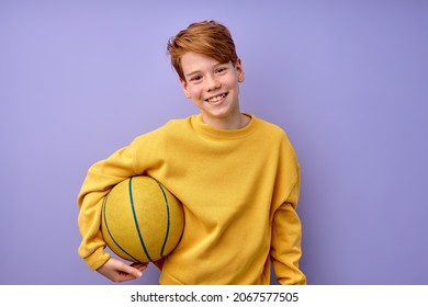 Happy teenage boy in yellow shirt holding basketball ball in hands isolated in studio on purple background, portrait. pozitive excited kid looking at camera, laughing, sport, childhood concept