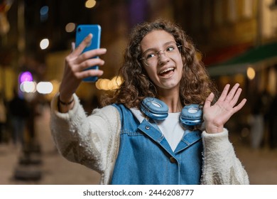Happy teenage child girl blogger taking selfie on smartphone communicating video call online with subscribers recording stories for social media vlog. Young woman standing on city street at evening