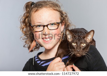 Happy teenage girl with glasses and blond curly hair hugging dark brown eastern shorthair cat. Studio shot isolated on grey background.