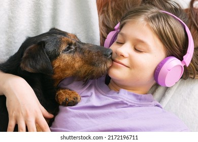 Happy teenage girl in lilac t-shirt in pink headphones listens to music and has fun playing with dog - Powered by Shutterstock