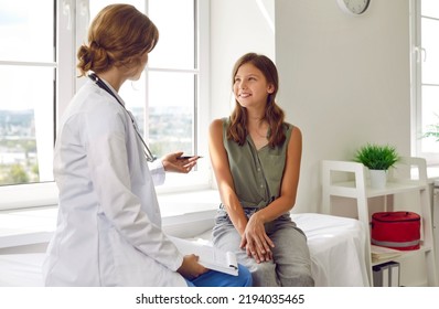 Happy teenage girl is listening to doctor during medical examination in modern clinic. Caucasian girl sits on examination couch in exam room and listens to advice of female pediatrician.
