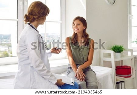 Happy teenage girl is listening to doctor during medical examination in modern clinic. Caucasian girl sits on examination couch in exam room and listens to advice of female pediatrician.