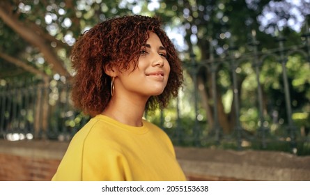 Happy teenage girl looking away with a smile outdoors. Cheerful female youngster wearing red hair and a mustard sweatshirt in the city. Young teenager standing alone on an urban sidewalk. - Shutterstock ID 2055153638