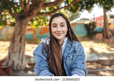 Happy Teenage Girl Looking At The Camera With A Smile. Cheerful Brunette Girl Wearing A Denim Jacket Outdoors In The City. Female Youngster Sitting Alone In An Urban Park During The Day.