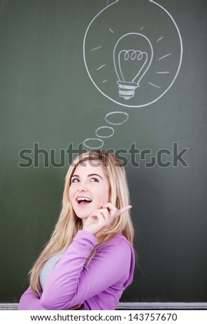 Happy teenage girl looking up while standing next to bulb drawn in thought bubble against chalkboard