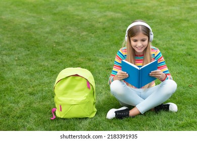 Happy teenage girl reading and listening to audiobook in headphones sitting on grass after school - Powered by Shutterstock