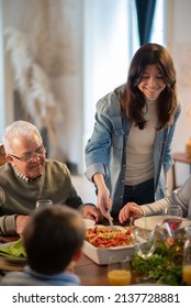 Happy Teenage Girl Serving Pasta At Family Dinner. Multigenerational Family Sitting At Table During Party. Family Party Concept