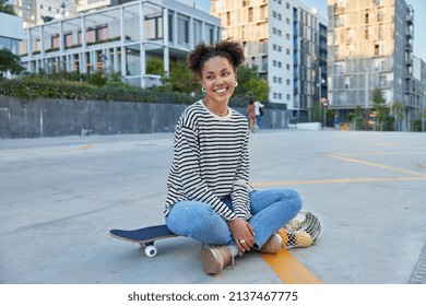 Happy teenage girl sits crossed legs on longboard rests after riding smiles joyfully poses in urban setting during daytime against blurred background. Youth lifestyle and recreation concept. - Powered by Shutterstock