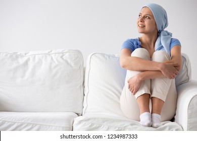 Happy teenage girl suffering from lung cancer, wearing blue headscarf and siting on the couch at hospice