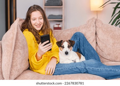 Happy teenage girl wearing yellow hoodie looks at phone screen. Young woman with dog sitting at home on couch watching the news on her cellphone, communicating in social networks. Generation Z - Powered by Shutterstock