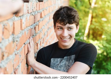 A happy teenage guy rests in nature near an old brick house and smiles sincerely and looks away, happy moments of a teenager's life, the boy has beautiful white teeth and dark hair - Shutterstock ID 2200452407