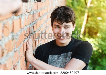 A happy teenage guy rests in nature near an old brick house and smiles sincerely and looks away, happy moments of a teenager's life, the boy has beautiful white teeth and dark hair