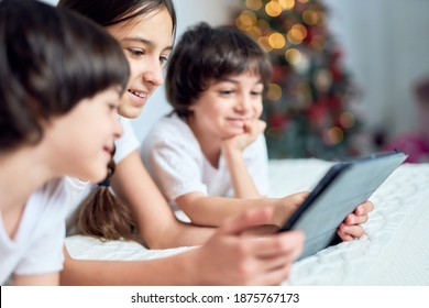 Happy teenage latin girl smiling while spending time with her siblings, using digital tablet, lying on the bed at home decorated for hristmas